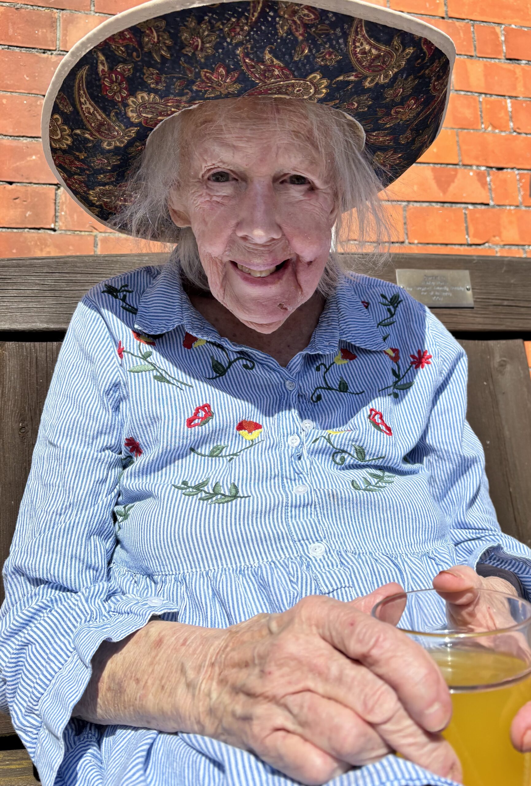 Female resident sat on a bench in the garden enjoying the sunshine with a glass of squash wearing a sun hat scaled