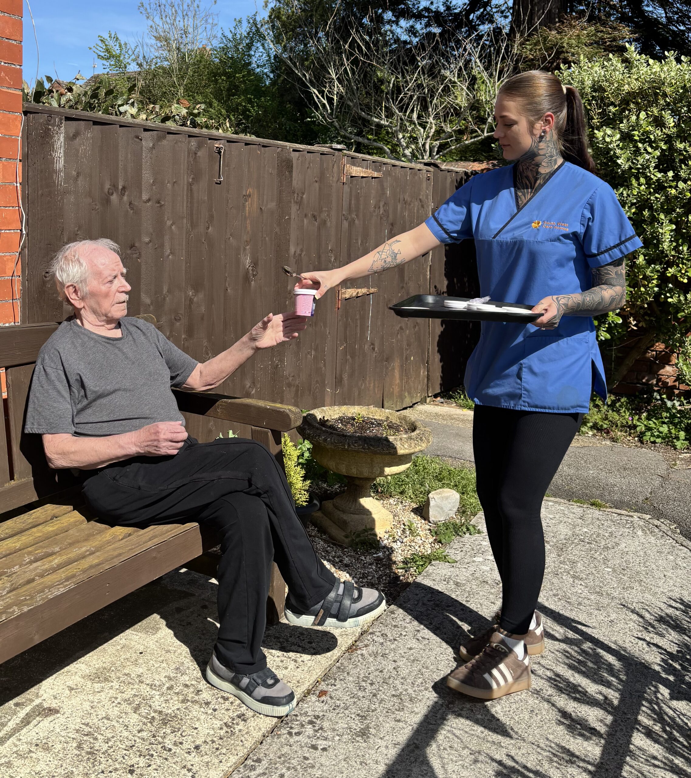 Male resident sat on a bench in the garden enjoying the sunshine with carer handing him a pot of ice cream scaled