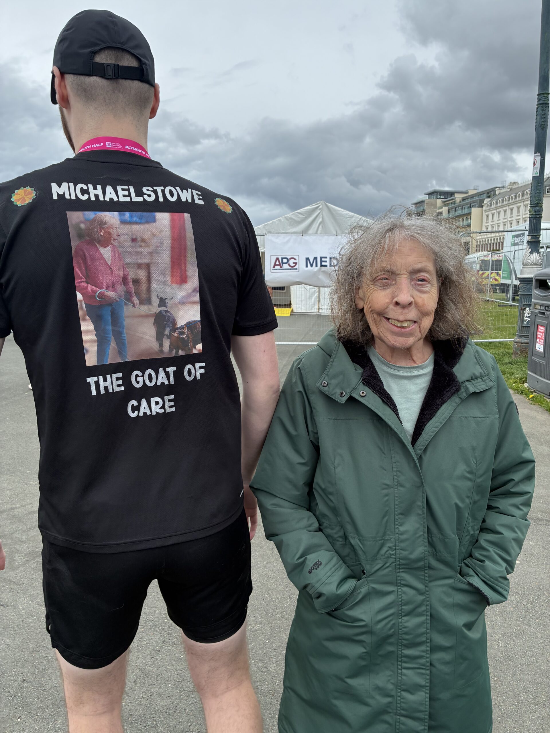 Carol stood beside Luke who is showing the back of his T-shirt which says Michaelstowe The GOAT of care