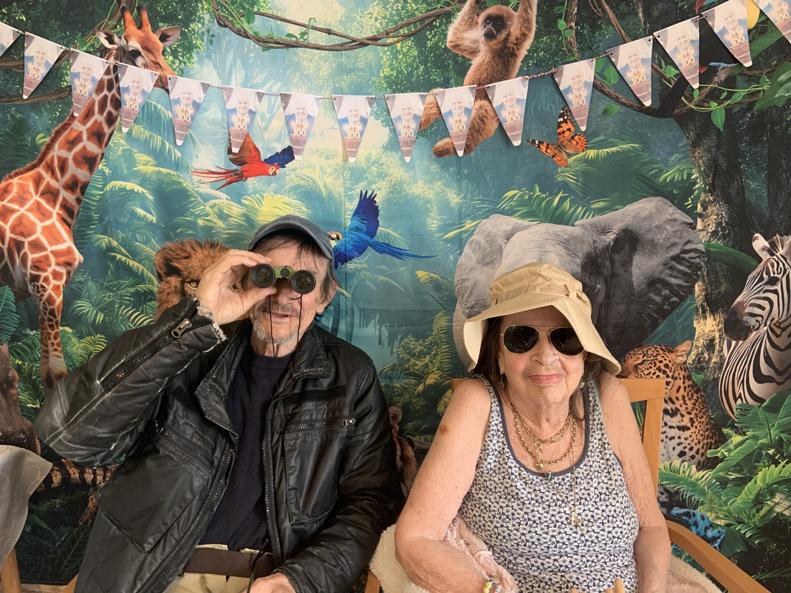 Male resident holding binoculars seated with a female resident in front of a jungle back drop