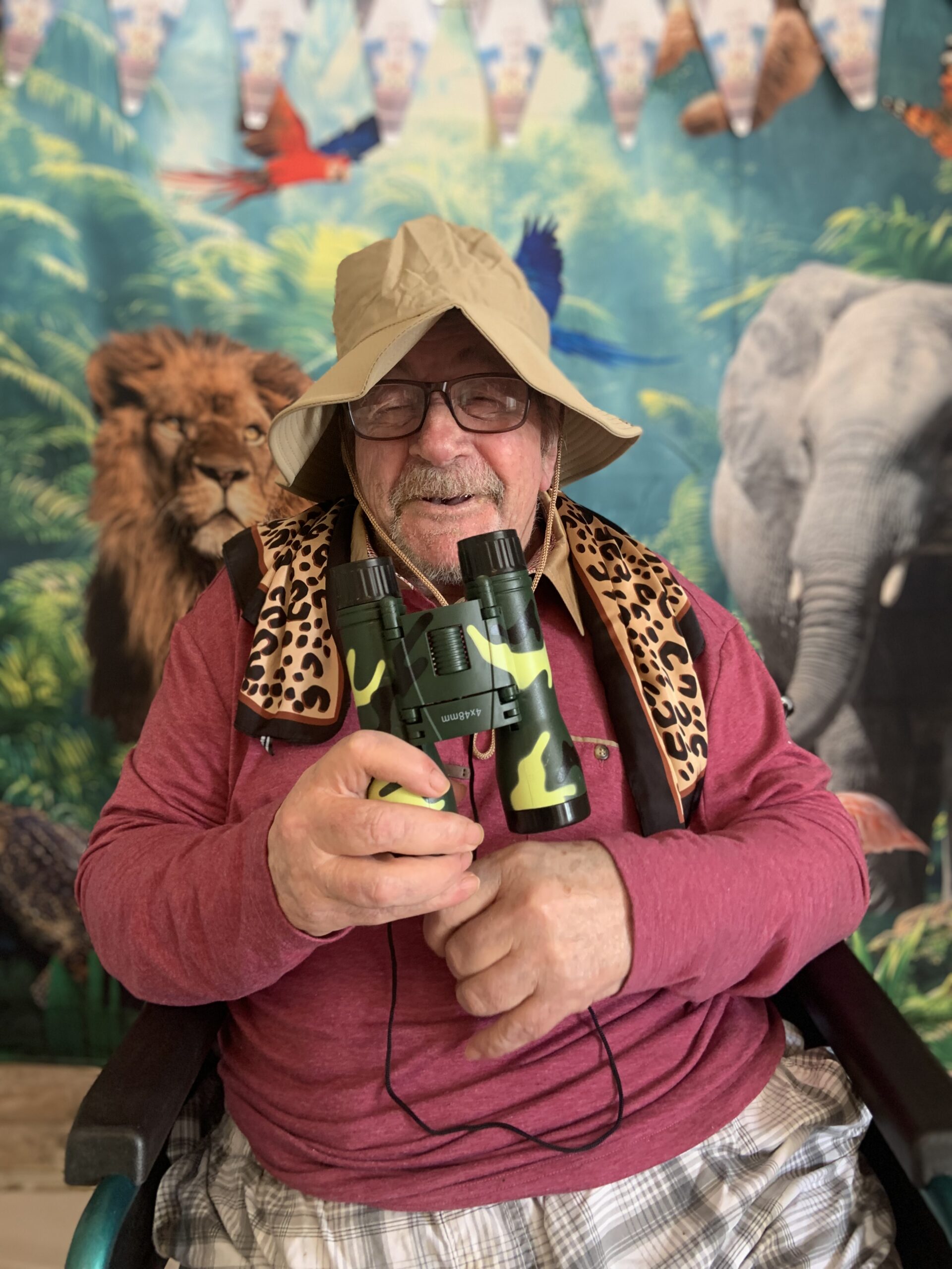 Male resident in a safari hat with binoculars seated in front of jungle back drop