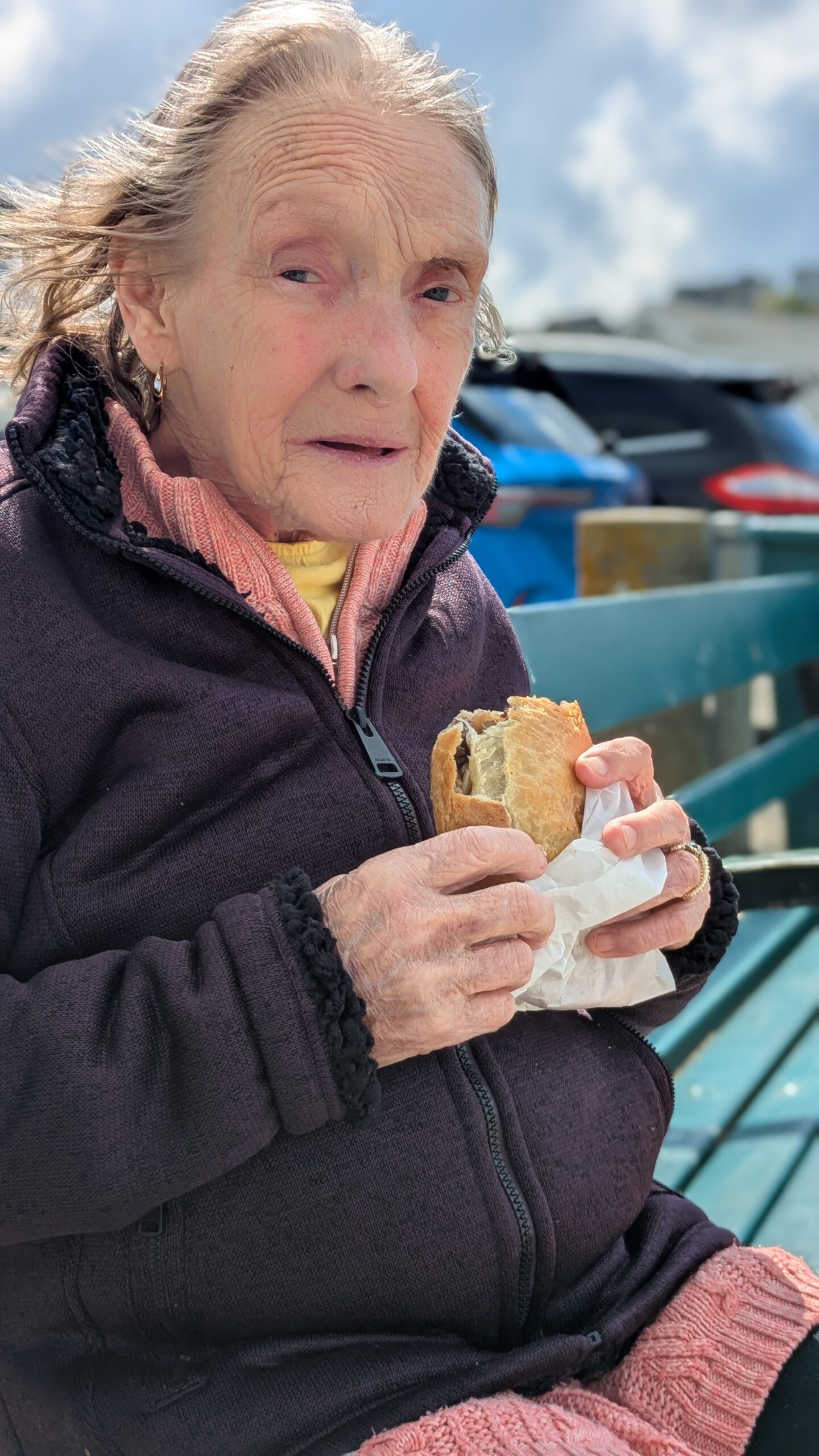 Thelma seated on a bench with a Cornish pasty enjoying the view