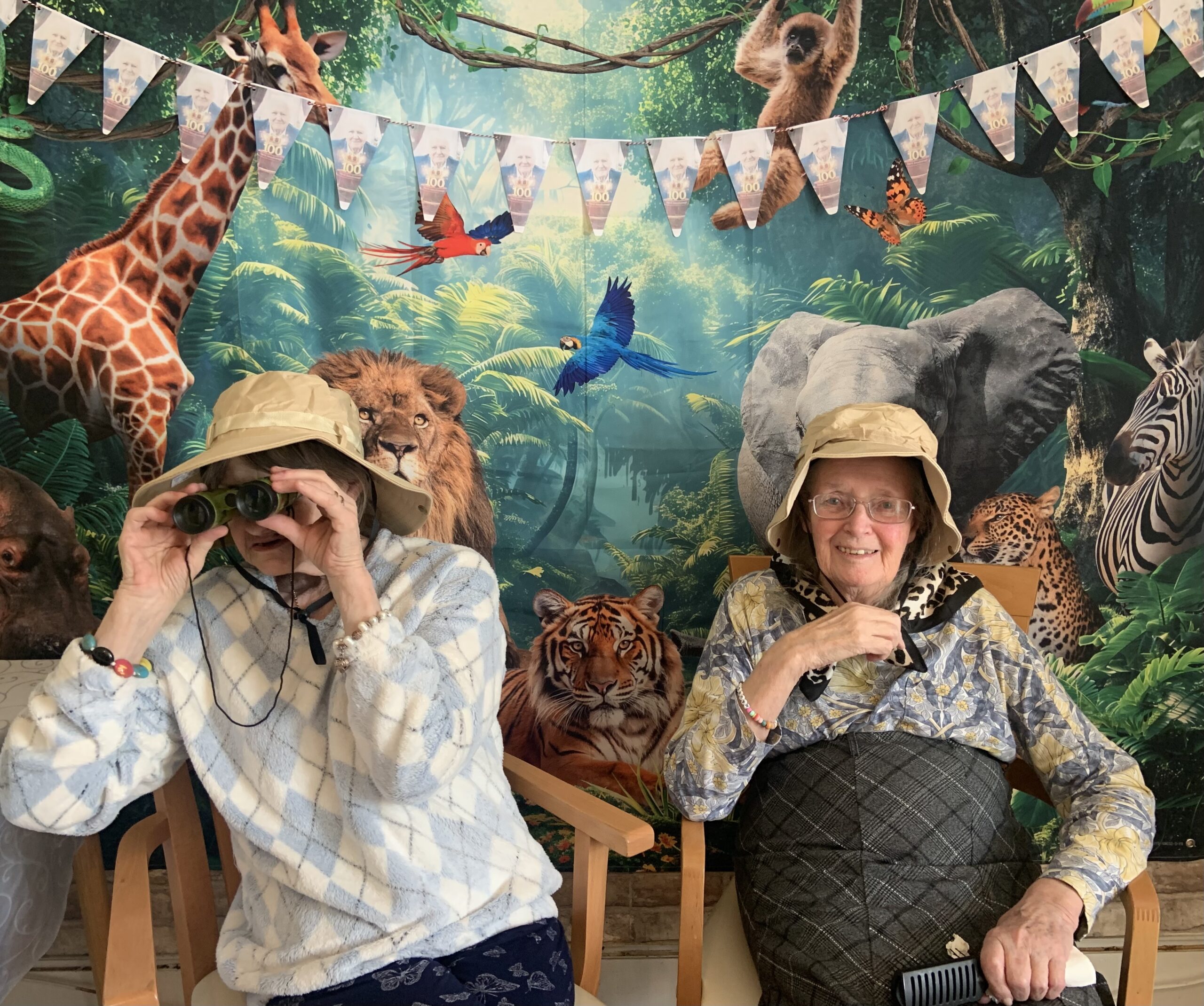 Two female residents in safari hats, one with binoculars seated in front of jungle back drop